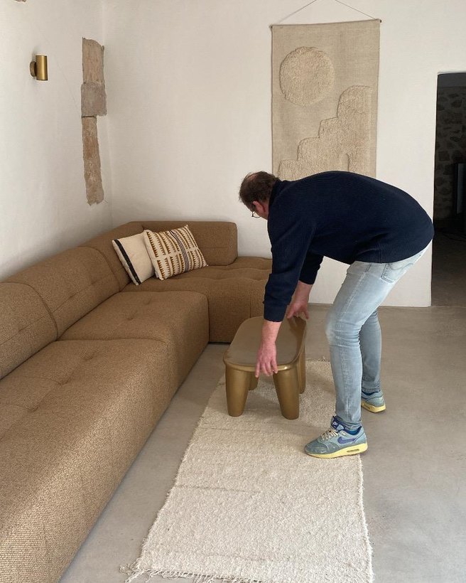 Iemand zet een laag meubelstuk op een wollen vloerkleed naast een brede bank, in een ruimte met houten balken